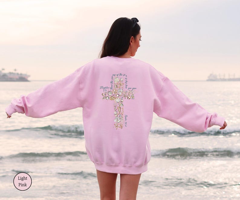 Puede incluir: Sudadera rosa claro con un dise&ntilde;o de cruz en la espalda. La cruz est&aacute; hecha de conchas marinas y elementos florales, con texto incorporado en el dise&ntilde;o. La sudadera es de color rosa claro liso.