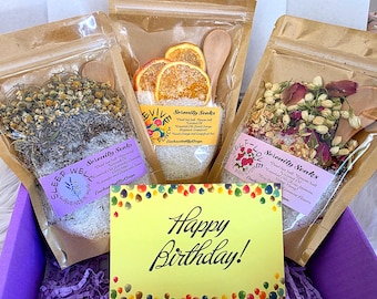 May include: Three clear pouches filled with bath salts, dried flowers, and wooden spoons. One pouch has orange slices, another has lavender, and the third has rose petals. A yellow "Happy Birthday!" card is in front.