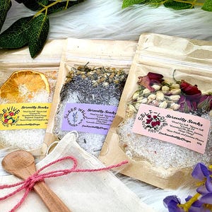 Pode incluir: Três sacos transparentes de sais de banho com flores e frutas secas. Cada saco tem um rótulo com texto. Uma colher de pau e um pequeno saco de tecido com uma corda rosa estão em primeiro plano. Os sacos estão rotulados como "Revive", "Sleep Well" e "Self-Love".