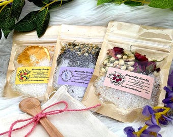 May include: Three clear bags of bath salts with dried flowers and fruit. Each bag has a label with text. A wooden spoon and small cloth bag with a pink string are in the foreground. The bags are labeled "Revive", "Sleep Well", and "Self-Love".