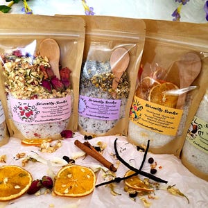 Può includere: Diversi sacchetti trasparenti pieni di sali da bagno, fiori secchi e cucchiai di legno. I sacchetti hanno etichette con la scritta "Serenity Soaks". Fette d'arancia, bastoncini di cannella e baccelli di vaniglia sono sparsi intorno ai sacchetti.