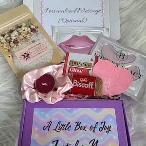 Peut inclure: Une boîte cadeau violette avec le texte "A Little Box of Joy Just for You" contient divers articles de bien-être. On y trouve des sels de bain, un masque pour les lèvres, des patchs pour les yeux, une bougie, un chouchou, une éponge en forme de cœur et un biscuit Lotus Biscoff.