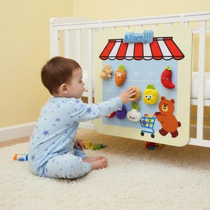 May include: A child interacts with a toy grocery store. The toy has a light yellow background with a red and white striped awning. It features plush vegetables and a bear with a shopping cart. The text "Allam!!!" is above the awning. The toy is attached to a cot.