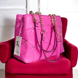 Puede incluir: Un bolso acolchado de color rosa brillante con correas de cadena doradas y tachuelas. El bolso tiene una forma estructurada y una etiqueta con un logotipo. El bolso está sobre una silla roja.
