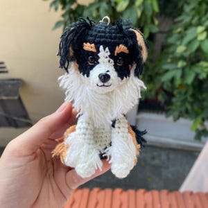 Könnte beinhalten: Handgefertigte Häkelfigur eines Hundes in sitzender Position. Der Hund hat ein Farbschema aus Schwarz, Weiß und Hellbraun. Der Hund hat eine flauschige weiße Brust und eine schwarze Nase. Die Figur wird in einer Hand gehalten.