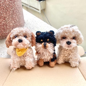 May include: Three handmade crochet dog figurines. The dogs have curly fur in shades of beige, brown, and black. One dog wears a yellow bandana. The dogs are sitting on a brown surface.