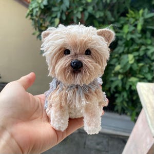 May include: A handcrafted Yorkshire Terrier dog ornament in shades of tan and cream. The dog features a textured, fluffy design with a black nose and dark eyes. The small figurine is held in a hand, highlighting its intricate details and size.