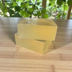 May include: Two rectangular bars of translucent, pale yellow soap are stacked on a wooden surface. The soap has a smooth, slightly glossy appearance. The background features green foliage.