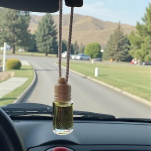 May include: A car air freshener with a clear glass bottle and a wooden cap, suspended by a beige rope. The bottle contains a yellow liquid. The background shows a road, trees, and a mountain range.