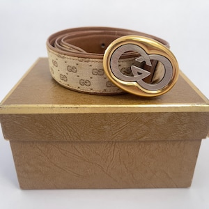 May include: A beige belt with a repeating brown logo pattern and a tan leather interior. The belt features a large, oval-shaped buckle with a gold-tone outer rim and a silver-tone interlocking G logo center, resting on a textured brown box.
