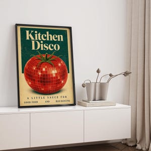 May include: A framed vertical poster featuring a red disco ball shaped like a tomato against a dark green background. The text reads Kitchen Disco at the top and A LITTLE SAUCE FOR GOOD FOOD AND BAD DANCING at the bottom in a vintage font.