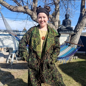 May include: A woman wearing a long, green and black patterned jacket with fringe. The jacket has a shawl collar and is open in the front. The woman is standing in a grassy area with a hammock and a statue of Buddha in the background.
