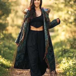 Può includere: Un cappotto lungo e fantasia con finiture a frange, indossato sopra un top corto e pantaloni neri. Il cappotto presenta un design verde acqua e marrone con cappuccio. L'outfit è completato da stivali neri, creando un'estetica bohémien.