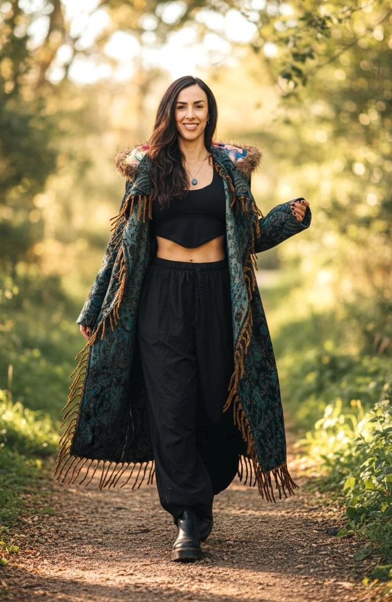 May include: A long, patterned coat with fringe trim, worn over a black crop top and pants. The coat features a teal and brown design with a hood. The outfit is completed with black boots, creating a bohemian aesthetic.