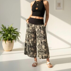 May include: Black halter top and wide-leg pants with a black and beige wave and fish pattern. The pants have a wide black waistband and pockets. The model is wearing gold jewelry and white sandals.