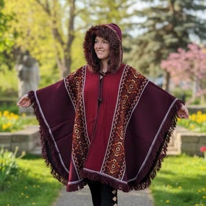 May include: A burgundy poncho with a hood trimmed with faux fur. The poncho features geometric patterns in shades of brown and cream, with fringe along the edges. A drawstring closure is visible at the neckline. The garment is designed for warmth and style.