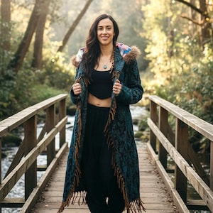 May include: A long, teal coat with a floral pattern and brown fringe is worn by a person standing on a wooden bridge. The coat has a hood with a colorful floral lining and faux fur trim. The person is wearing black pants and boots.