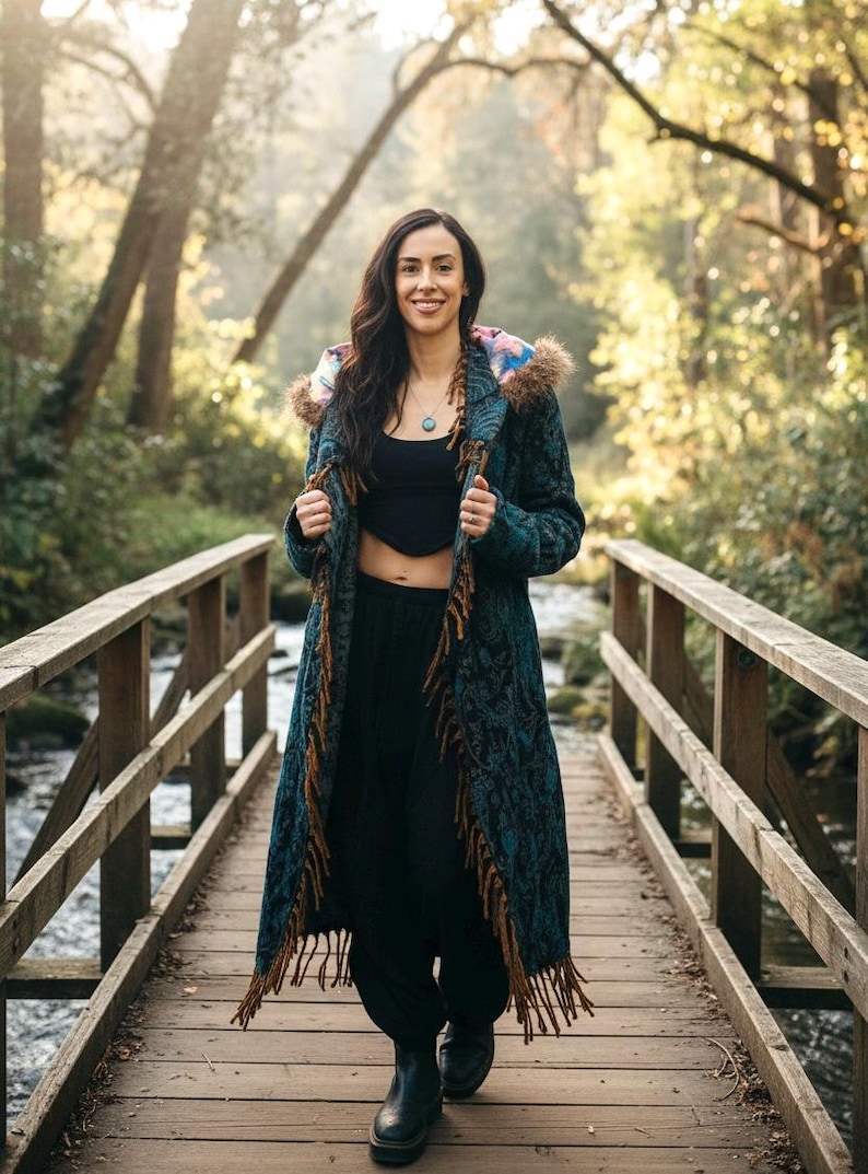May include: A long, teal coat with a floral pattern and brown fringe is worn by a person standing on a wooden bridge. The coat has a hood with a colorful floral lining and faux fur trim. The person is wearing black pants and boots.