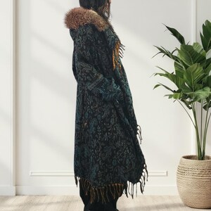 May include: A long, dark coat with a paisley pattern in teal and black. The coat features a faux fur-lined hood and fringe trim. The coat is worn over black pants and shoes.