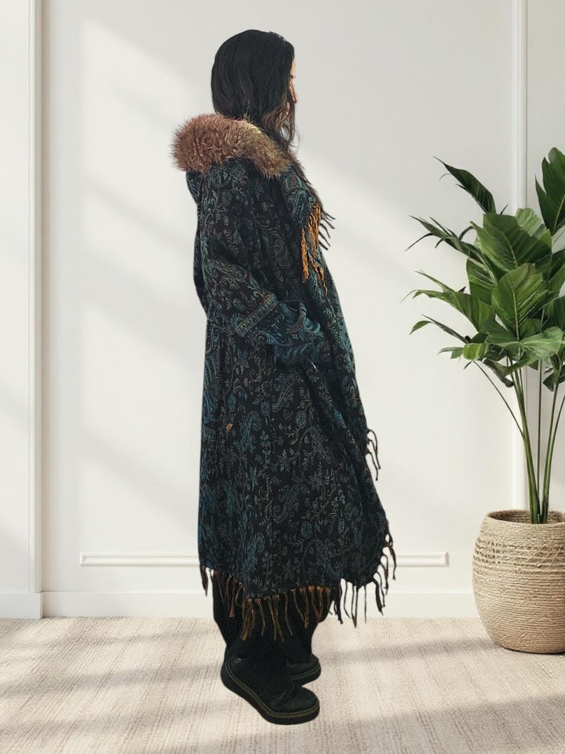 May include: A long, dark coat with a paisley pattern in teal and black. The coat features a faux fur-lined hood and fringe trim. The coat is worn over black pants and shoes.