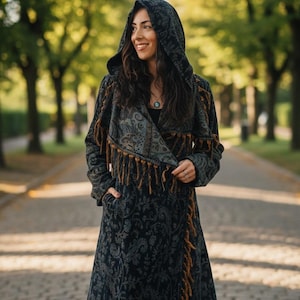 May include: A long, hooded coat with a paisley pattern in shades of black and grey. The coat features a wrap-style front with brown fringe trim along the edges. The coat is worn outdoors on a paved path lined with trees.