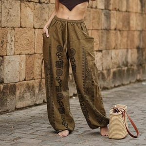 May include: Olive green harem pants with black patterned designs. The pants have a drawstring waist and pockets. A woven straw bag with a brown leather strap sits on the ground.