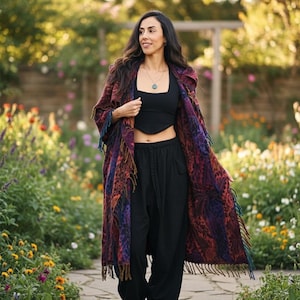 May include: A long, patterned coat with fringe in shades of red, purple, and blue. The coat is worn over a black top and black trousers. The person is walking on a stone path in a garden.