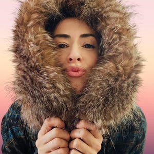 May include: A person wearing a brown fur-lined hood and a patterned jacket. The hood frames the face, with the fur adding texture and warmth. The jacket has a blue and black pattern. The person is holding the jacket closed.