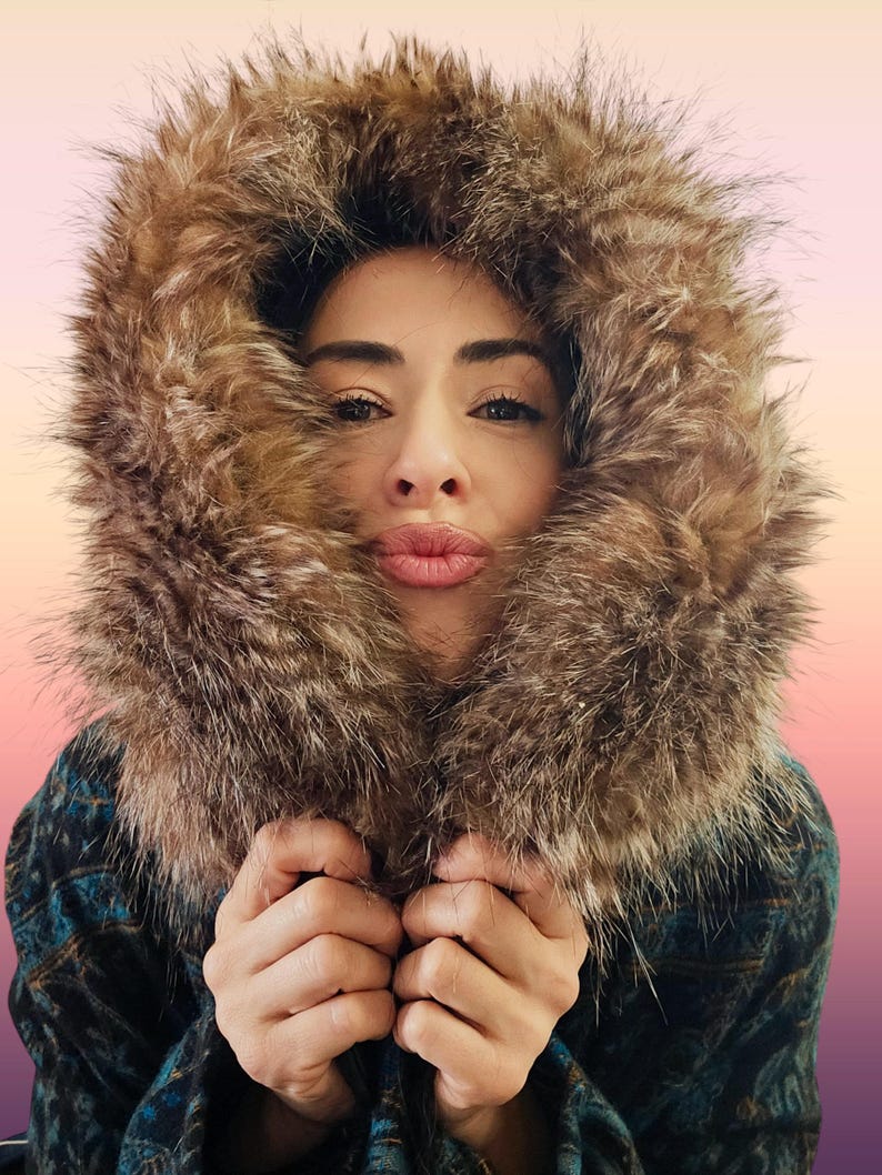 May include: A person wearing a brown fur-lined hood and a patterned jacket. The hood frames the face, with the fur adding texture and warmth. The jacket has a blue and black pattern. The person is holding the jacket closed.