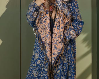 Puede incluir: Abrigo largo azul con estampado de cachemira y capucha forrada de piel sintética. El abrigo presenta un chal color melocotón con flecos y se lleva con leggings oscuros. El abrigo es de estilo bohemio.