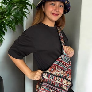 Puede incluir: Una persona lleva una camiseta negra y un sombrero de cubo multicolor con un patrón geométrico. También lleva una bolsa de bandolera multicolor con un patrón geométrico.