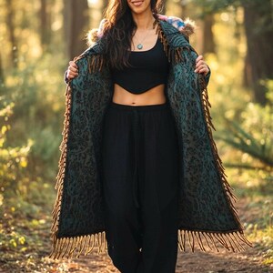 May include: A woman wearing a long, patterned coat with fringe, black top, and black pants. The coat is teal and brown with a floral design. The woman is smiling and standing in a forest.