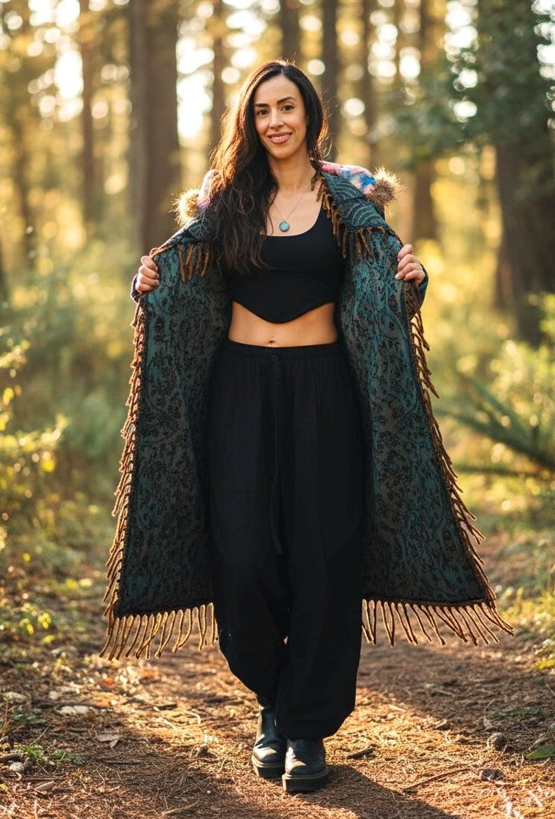 May include: A woman wearing a long, patterned coat with fringe, black top, and black pants. The coat is teal and brown with a floral design. The woman is smiling and standing in a forest.