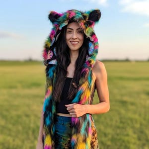 May include: A hooded scarf made of faux fur featuring a vibrant, multi-coloured tie-dye pattern in shades of blue, pink, yellow, and black. The hood includes two pointed animal-like ears at the top and long, dangling scarf ends on each side.