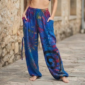May include: Blue tie-dye trousers with a relaxed fit. The trousers have a drawstring waist, pockets, and colourful abstract designs in red, yellow, and black. The fabric appears lightweight and comfortable.