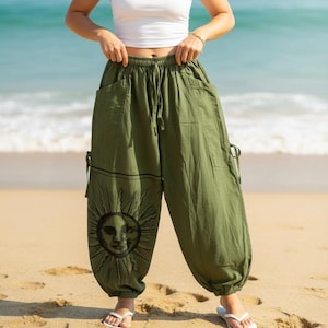 May include: Olive green loose-fitting trousers featuring an elastic waistband with a drawstring, side pockets, and a black graphic print of a sun and moon face on the lower left leg. The pants have a relaxed silhouette with gathered ankles.
