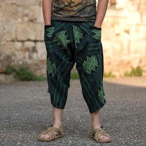 May include: Cropped trousers in a dark green and black color palette featuring a geometric diamond pattern. These loose-fitting pants have a drop-crotch design, side pockets, and a wide waistband, paired with beige woven rope sandals.