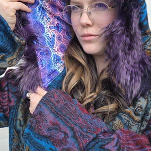 May include: A hooded coat with purple faux fur trim and lining. The coat has a colourful pattern in shades of blue, purple, and brown. The hood is up, displaying the intricate design of the lining.
