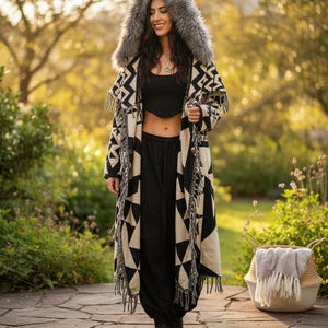 May include: A long, open-front coat featuring a black and cream geometric pattern, a large hood with grey faux fur trim, and fringe detailing along the edges. The coat is worn over a black cropped tank top and black trousers with black boots.