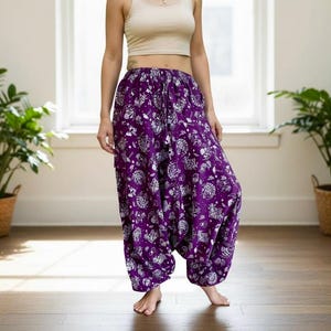 May include: Purple harem pants with a white floral pattern. The pants have a loose, comfortable fit and a drawstring waist. The model is wearing a beige crop top.