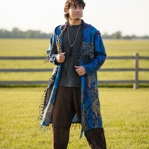 May include: A long, hooded coat in shades of blue and tan with a textured, patterned fabric and fringe trim along the edges. The coat is worn over a dark grey t-shirt and loose-fitting brown trousers, paired with a long beaded necklace with a tassel.