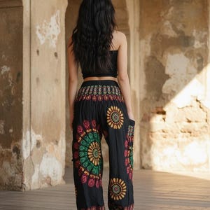 Puede incluir: Pantalones har&eacute;n negros con un vibrante estampado floral en rojo, verde y amarillo. Los pantalones tienen un ajuste c&oacute;modo y holgado y se usan con un top corto negro. El fondo es una estructura de piedra iluminada por el sol.