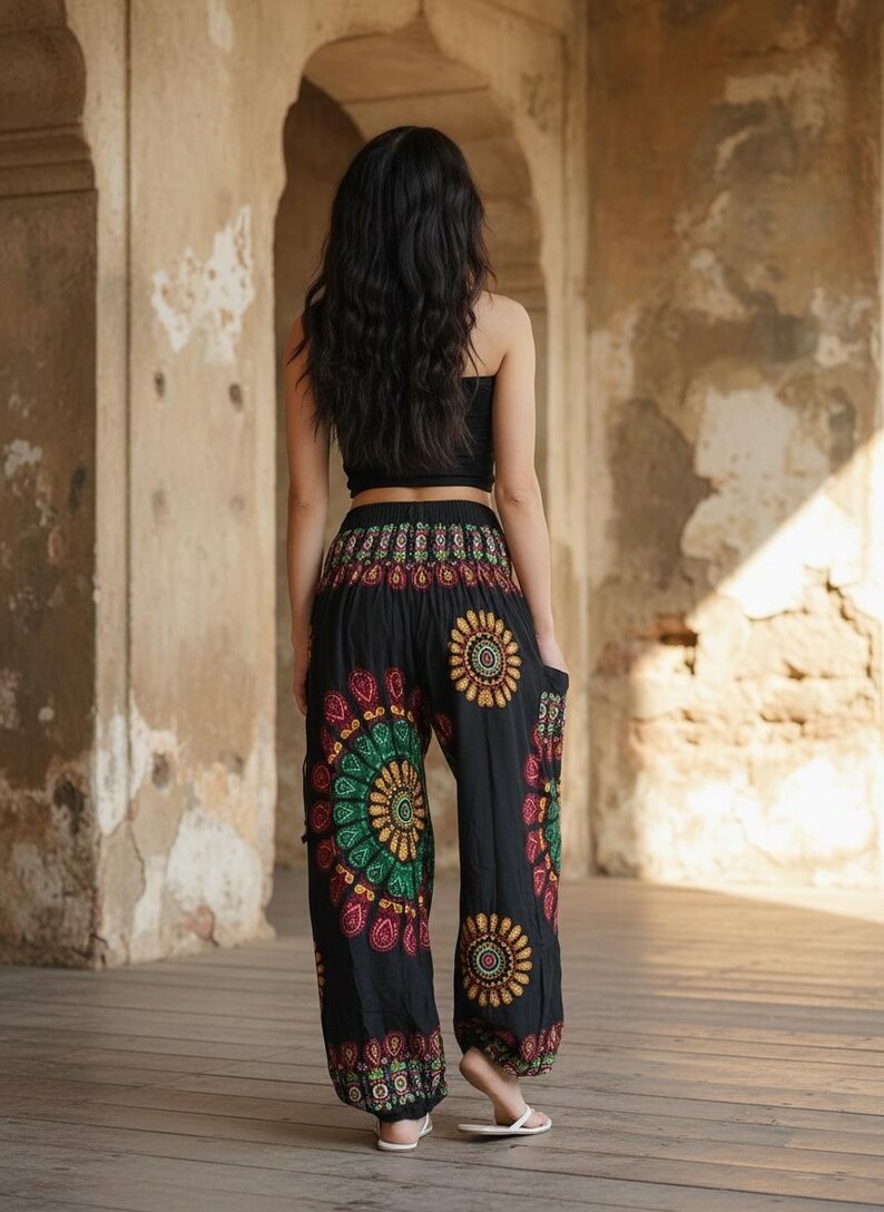 Puede incluir: Pantalones har&eacute;n negros con un vibrante estampado floral en rojo, verde y amarillo. Los pantalones tienen un ajuste c&oacute;modo y holgado y se usan con un top corto negro. El fondo es una estructura de piedra iluminada por el sol.