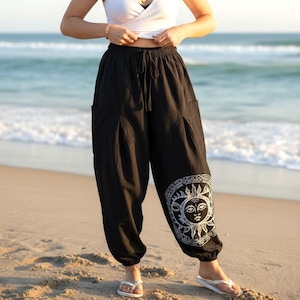 May include: Black loose-fitting pants with an elastic waistband and a drawstring tie. The pants feature a white graphic print of a sun and moon design on the lower left leg, side pockets, and gathered elastic cuffs at the ankles.