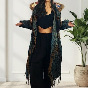 May include: A long, patterned coat with fringe and a fur-trimmed hood. The coat features a teal and brown color scheme with intricate designs. The model is wearing black pants and a black top. The coat is a statement piece.