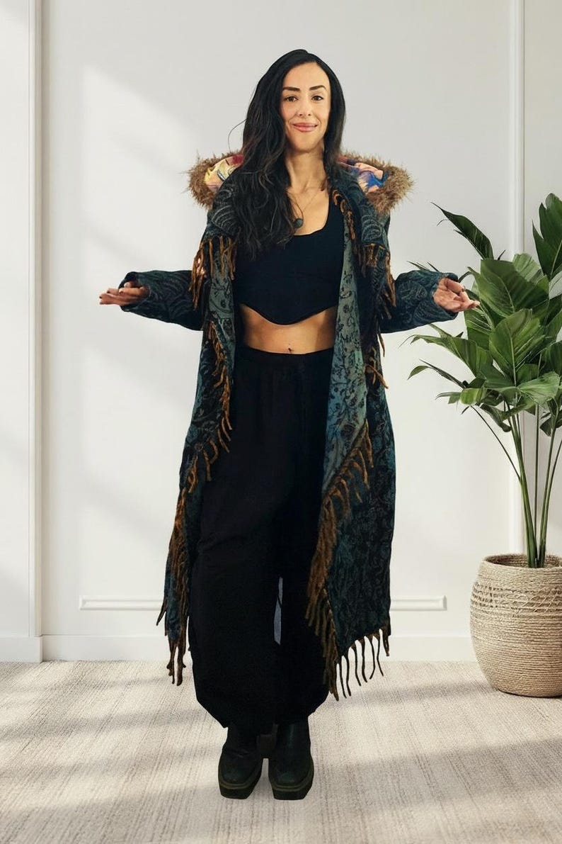 May include: A long, patterned coat with fringe and a fur-trimmed hood. The coat features a teal and brown color scheme with intricate designs. The model is wearing black pants and a black top. The coat is a statement piece.