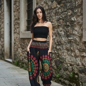 Puede incluir: Una persona con un top negro y pantalones harem negros con un dise&ntilde;o de mandala colorido. Los pantalones tienen bolsillos y se combinan con sandalias blancas. El fondo es una pared de piedra y un camino pavimentado.