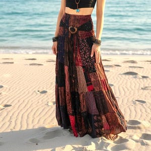 May include: A long, patchwork skirt in shades of red, brown, and black. The skirt features a wide belt with a wooden buckle. The skirt is worn on a sandy beach with the ocean in the background.