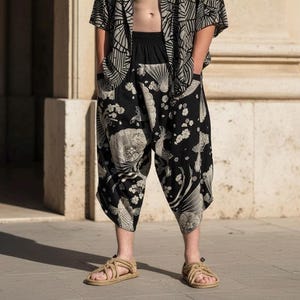 May include: Black drop-crotch pants featuring a white graphic print of koi fish, fans, and cherry blossoms. The pants have an elastic waistband and a loose, wide-leg silhouette that falls below the knees. The fabric is black with white patterns.