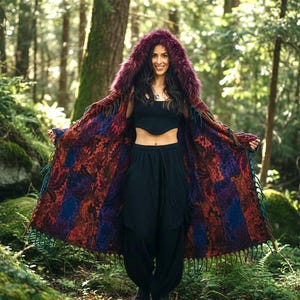 May include: A hooded cloak with a faux fur trim in shades of burgundy and purple. The cloak features a patterned design in red, blue, and purple, with fringe along the edges. The person is wearing black pants and a black top.